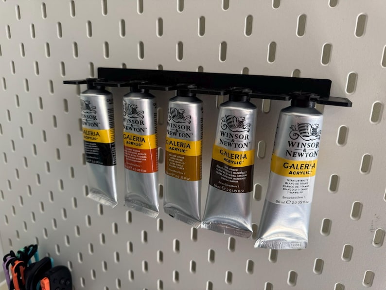May include: Five tubes of Winsor & Newton Galeria acrylic paint in various colours are displayed on a black metal rack. The tubes are silver with yellow and black labels, mounted on a white pegboard.