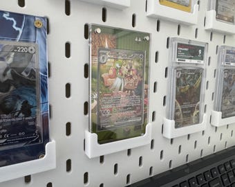 Puede incluir: Un tablero de clavijas blanco con varios soportes de tarjetas de plástico transparente que muestran tarjetas coleccionables. Las tarjetas están protegidas por fundas de plástico duro. Las tarjetas presentan ilustraciones de personajes y texto. El tablero de clavijas está montado sobre una superficie negra.