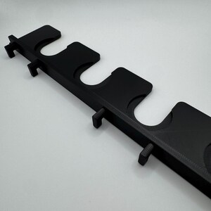 May include: A black, 3D-printed wall-mounted rack with multiple U-shaped slots and small protruding pegs. The rack is designed to hold items, possibly fishing rods or tools. The design is simple and functional, with a matte finish.