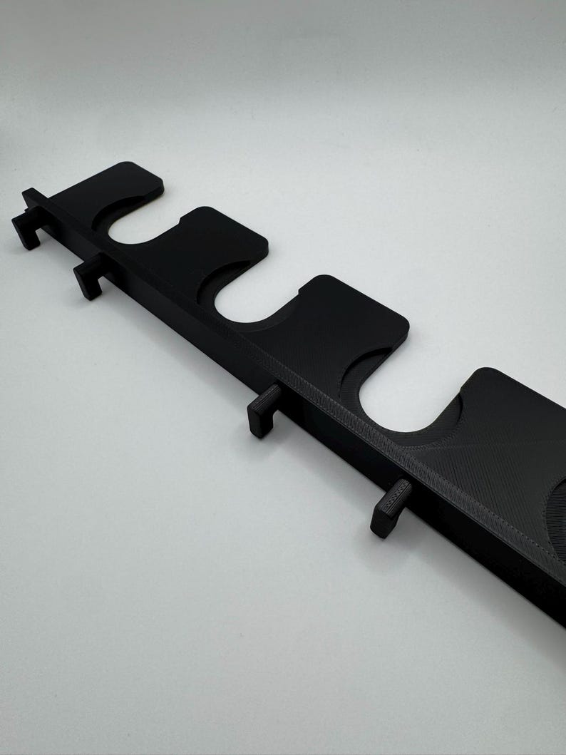 May include: A black, 3D-printed wall-mounted rack with multiple U-shaped slots and small protruding pegs. The rack is designed to hold items, possibly fishing rods or tools. The design is simple and functional, with a matte finish.