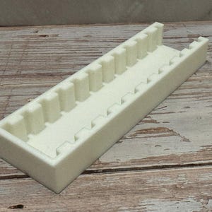 Puede incluir: Un organizador rectangular blanco roto, impreso en 3D, con una serie de divisores verticales y bordes dentados. El organizador est&aacute; dise&ntilde;ado para guardar objetos peque&ntilde;os y est&aacute; hecho de un material opaco s&oacute;lido. El art&iacute;culo est&aacute; sobre una superficie de madera.