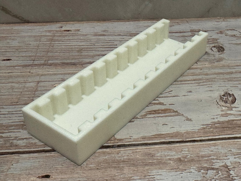 Puede incluir: Un organizador rectangular blanco roto, impreso en 3D, con una serie de divisores verticales y bordes dentados. El organizador est&aacute; dise&ntilde;ado para guardar objetos peque&ntilde;os y est&aacute; hecho de un material opaco s&oacute;lido. El art&iacute;culo est&aacute; sobre una superficie de madera.