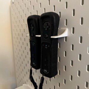 May include: Two black Wii remotes are mounted on a white pegboard using white holders. The remotes have black wrist straps and feature buttons and the Wii logo. The pegboard has evenly spaced holes.