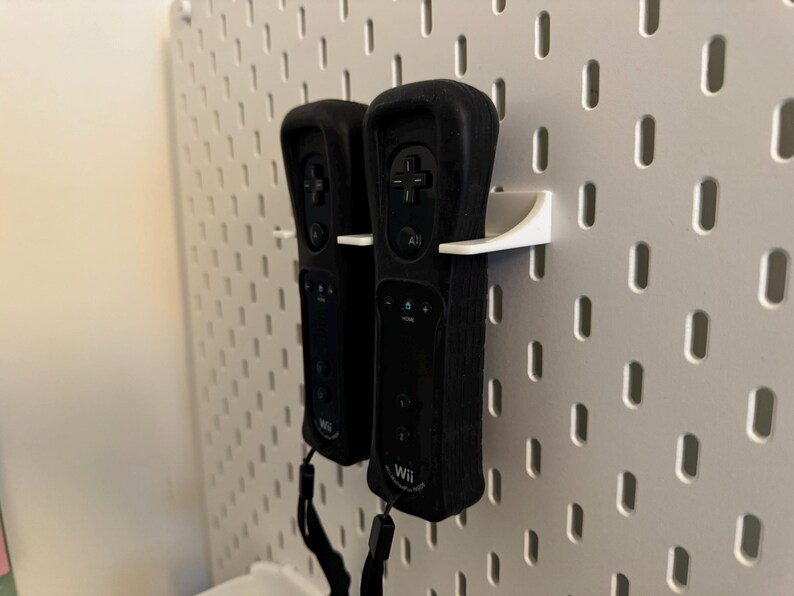 May include: Two black Wii remotes are mounted on a white pegboard using white holders. The remotes have black wrist straps and feature buttons and the Wii logo. The pegboard has evenly spaced holes.