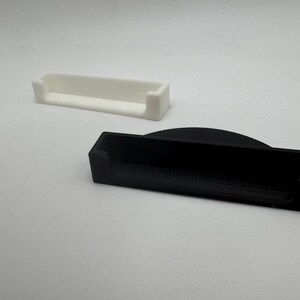 May include: Two rectangular trays, one white and one black, are displayed against a white background. The trays have a low-profile design with raised edges on the ends. The black tray has a curved top edge.