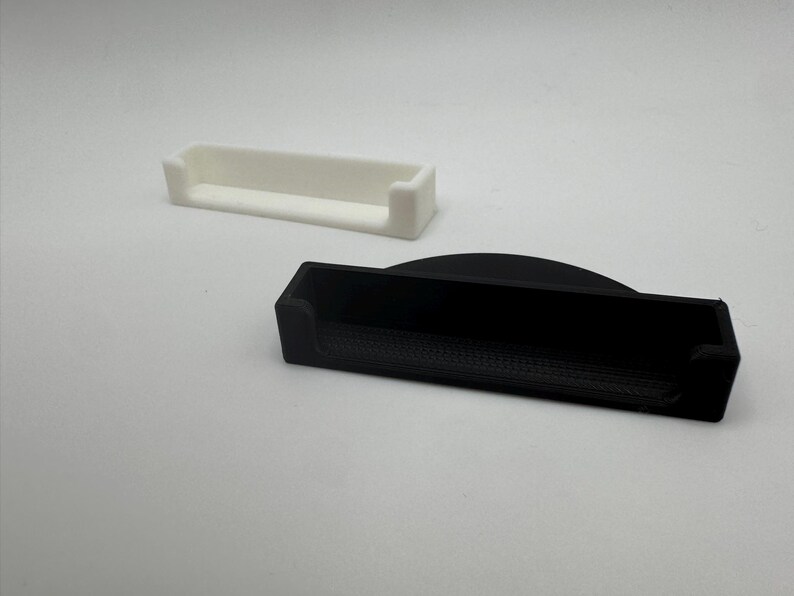 May include: Two rectangular trays, one white and one black, are displayed against a white background. The trays have a low-profile design with raised edges on the ends. The black tray has a curved top edge.