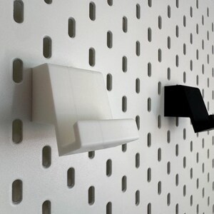 Op de afbeelding: Twee telefoonhouders, &eacute;&eacute;n wit en &eacute;&eacute;n zwart, gemonteerd op een wit pegboard. De houders zijn schuin en ontworpen om een telefoon vast te houden. Het pegboard heeft gelijkmatig verdeelde, ovale gaten.
