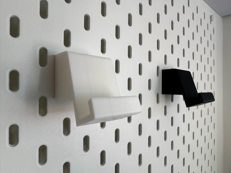 Op de afbeelding: Twee telefoonhouders, &eacute;&eacute;n wit en &eacute;&eacute;n zwart, gemonteerd op een wit pegboard. De houders zijn schuin en ontworpen om een telefoon vast te houden. Het pegboard heeft gelijkmatig verdeelde, ovale gaten.