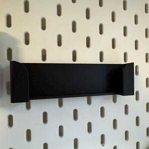 Puede incluir: Un estante negro con soportes laterales est&aacute; montado en una pared perforada blanca. El estante es rectangular y parece estar hecho de metal o un material similar. La pared perforada tiene agujeros ovalados espaciados.