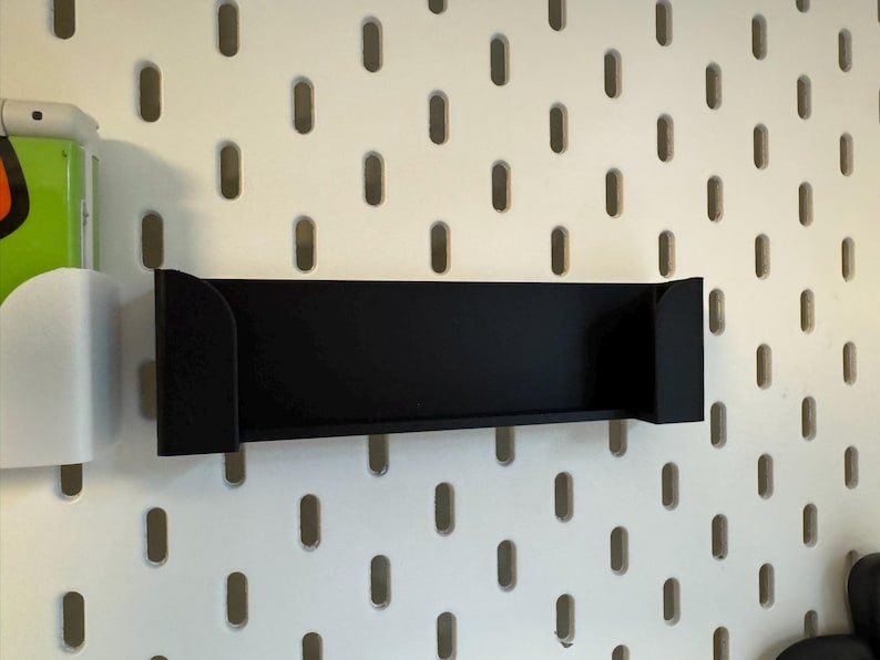 Puede incluir: Un estante negro con soportes laterales est&aacute; montado en una pared perforada blanca. El estante es rectangular y parece estar hecho de metal o un material similar. La pared perforada tiene agujeros ovalados espaciados.