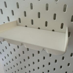 May include: White plastic shelf with a curved front edge, designed to hang on a pegboard wall.