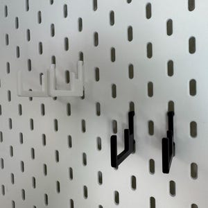 May include: White and black pegboard hooks. Two white hooks are positioned at the top, and two black hooks are below. The hooks are designed to fit into the oval-shaped holes of a pegboard. The pegboard is white.