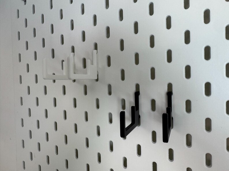 May include: White and black pegboard hooks. Two white hooks are positioned at the top, and two black hooks are below. The hooks are designed to fit into the oval-shaped holes of a pegboard. The pegboard is white.