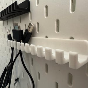May include: White pegboard with black and white cable organizers. The organizers are designed to hold various types of cables, with multiple slots for each cable. The pegboard has evenly spaced holes.