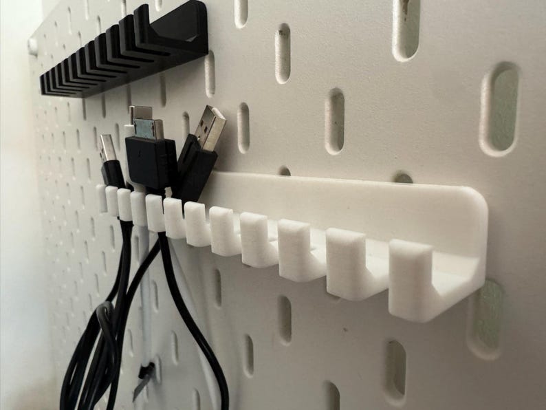May include: White pegboard with black and white cable organizers. The organizers are designed to hold various types of cables, with multiple slots for each cable. The pegboard has evenly spaced holes.