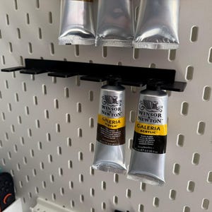 May include: A black metal paint tube holder with several tubes of Winsor & Newton Galeria acrylic paint. The tubes are silver with yellow and black labels. The holder is mounted on a white pegboard.