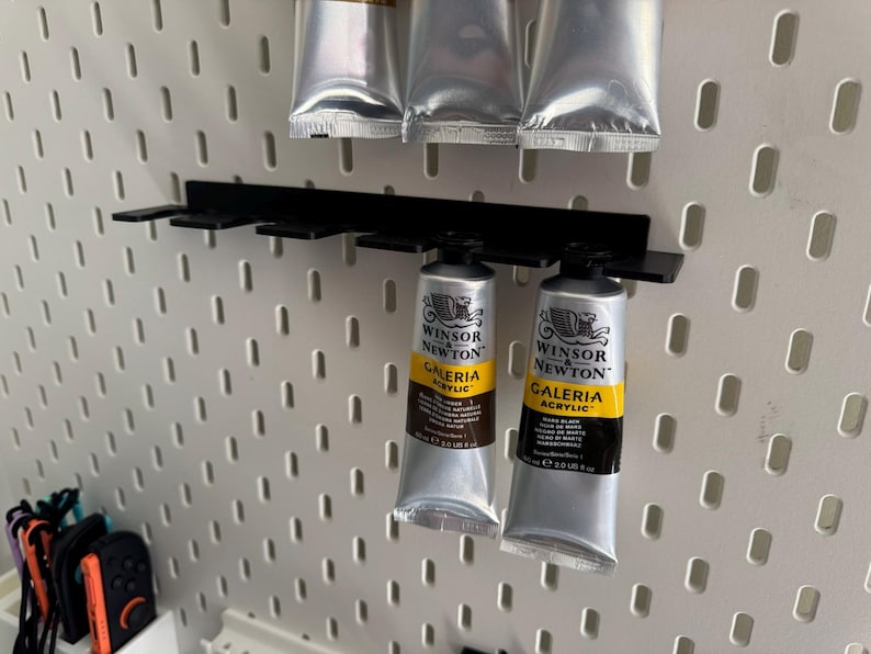 May include: A black metal paint tube holder with several tubes of Winsor & Newton Galeria acrylic paint. The tubes are silver with yellow and black labels. The holder is mounted on a white pegboard.
