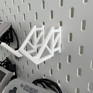 Puede incluir: Soporte de pared blanco impreso en 3D para mandos de videojuegos. El soporte est&aacute; dise&ntilde;ado para sujetar dos mandos y tiene forma triangular. El soporte est&aacute; fijado a una pared de tablero perforado.