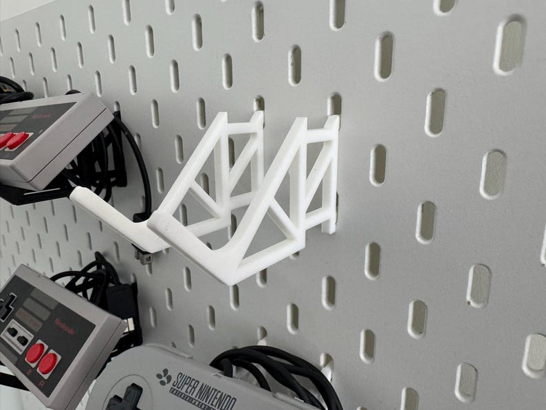 Puede incluir: Soporte de pared blanco impreso en 3D para mandos de videojuegos. El soporte est&aacute; dise&ntilde;ado para sujetar dos mandos y tiene forma triangular. El soporte est&aacute; fijado a una pared de tablero perforado.