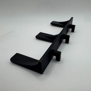 May include: A black, 3D-printed shelf with three angled supports. The shelf is designed to hold items and has a minimalist aesthetic. The supports are evenly spaced along the shelf's length.