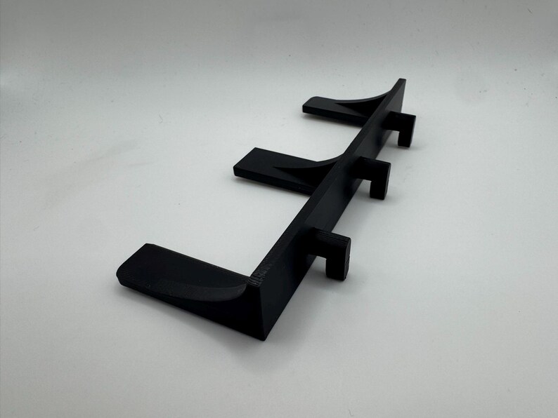 May include: A black, 3D-printed shelf with three angled supports. The shelf is designed to hold items and has a minimalist aesthetic. The supports are evenly spaced along the shelf's length.