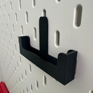 May include: Black plastic pegboard hook with a small shelf. The hook is mounted on a white pegboard.