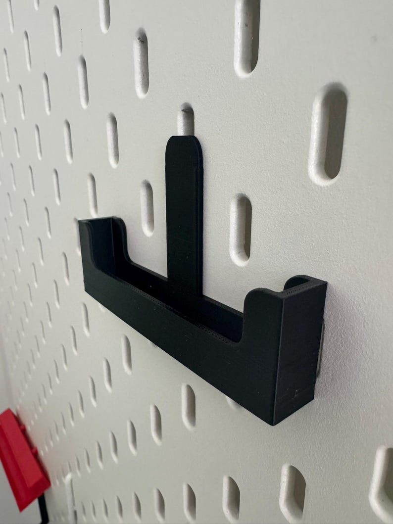May include: Black plastic pegboard hook with a small shelf. The hook is mounted on a white pegboard.