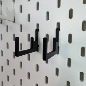 May include: Two black pegboard hooks are displayed against a white pegboard background. The hooks are made of a sturdy material and are designed to hold items. The pegboard has evenly spaced oval holes.