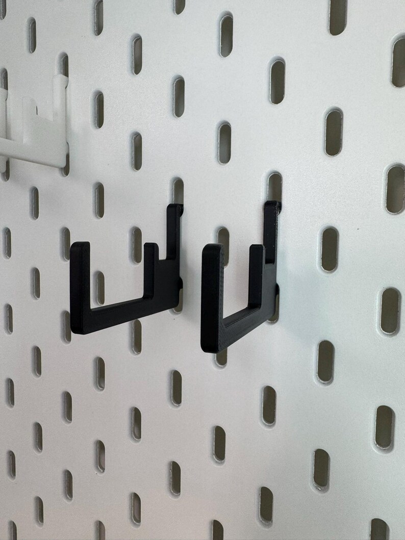 May include: Two black pegboard hooks are displayed against a white pegboard background. The hooks are made of a sturdy material and are designed to hold items. The pegboard has evenly spaced oval holes.