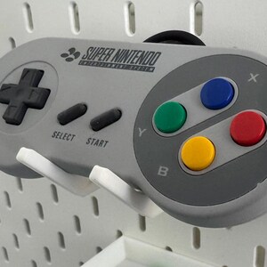 Puede incluir: Un mando de Super Nintendo Entertainment System gris con un pad direccional negro, cuatro botones de colores y el texto "SUPER NINTENDO ENTERTAINMENT SYSTEM".