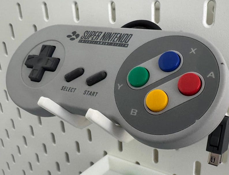 Puede incluir: Un mando de Super Nintendo Entertainment System gris con un pad direccional negro, cuatro botones de colores y el texto "SUPER NINTENDO ENTERTAINMENT SYSTEM".
