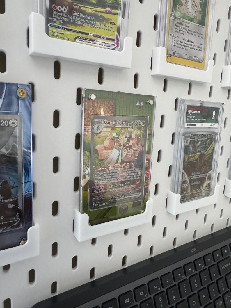K&ouml;nnte beinhalten: Eine wei&szlig;e Lochwand mit mehreren durchsichtigen Plastikkartenhaltern, die Sammelkarten enthalten. Die Karten zeigen Illustrationen von Pok&eacute;mon-Charakteren und haben Text in Englisch und Japanisch. Eine Tastatur ist teilweise im unteren rechten Eck des Bildes zu sehen.
