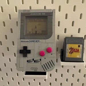 May include: A gray Nintendo Game Boy console with a game cartridge. The Game Boy has a screen displaying a game scene, a black directional pad, and pink buttons. The game cartridge features a Zelda game.