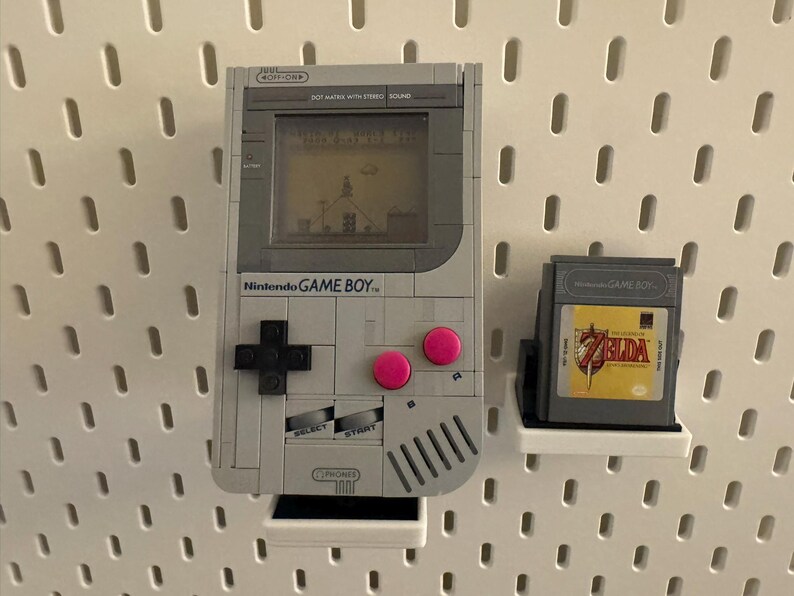 May include: A gray Nintendo Game Boy console with a game cartridge. The Game Boy has a screen displaying a game scene, a black directional pad, and pink buttons. The game cartridge features a Zelda game.