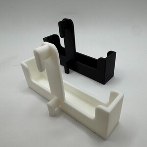 以下が含まれることがあります： 白と黒の2つの3Dプリントホルダーが白い背景に表示されています。各ホルダーは長方形のベースと上方に伸びる湾曲したフックを備えています。白いホルダーが手前にあり、黒いホルダーがその背後にあります。