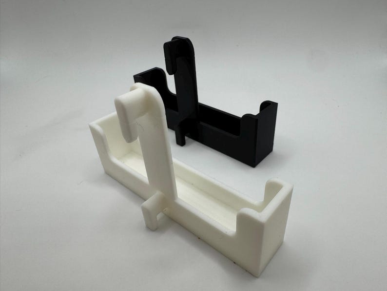 以下が含まれることがあります： 白と黒の2つの3Dプリントホルダーが白い背景に表示されています。各ホルダーは長方形のベースと上方に伸びる湾曲したフックを備えています。白いホルダーが手前にあり、黒いホルダーがその背後にあります。