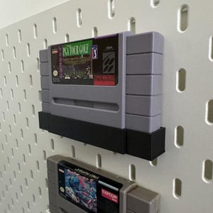K&ouml;nnte beinhalten: Zwei graue Super Nintendo-Spielmodule, eines mit der Aufschrift "PGA TOUR GOLF", an einer wei&szlig;en Lochwand befestigt. Die Module werden von schwarzen Halterungen gehalten. Das zweite Modul ist teilweise sichtbar, mit dem Text "STREET FIGHTER".