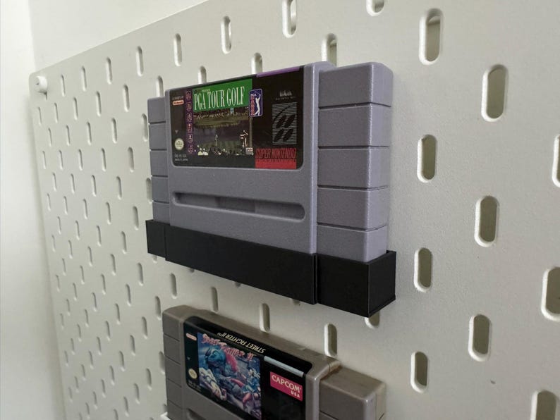 K&ouml;nnte beinhalten: Zwei graue Super Nintendo-Spielmodule, eines mit der Aufschrift "PGA TOUR GOLF", an einer wei&szlig;en Lochwand befestigt. Die Module werden von schwarzen Halterungen gehalten. Das zweite Modul ist teilweise sichtbar, mit dem Text "STREET FIGHTER".