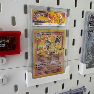 Peut inclure: Une carte Pok&eacute;mon Charizard class&eacute;e de la collection classique 2021 Celebrations, class&eacute;e 9 Mint. La carte est dans un &eacute;tui de protection transparent et expos&eacute;e sur un panneau perfor&eacute; blanc. La carte pr&eacute;sente une illustration de Charizard.
