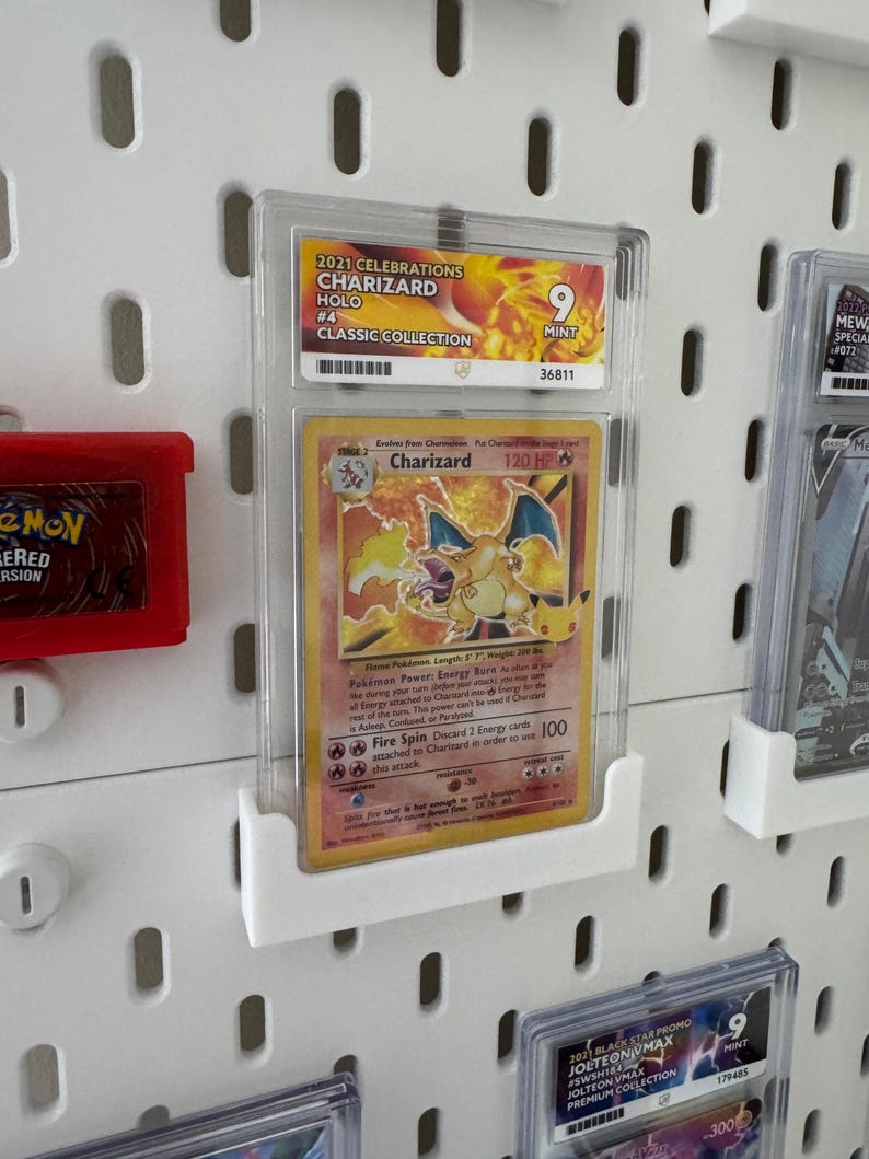 Peut inclure: Une carte Pok&eacute;mon Charizard class&eacute;e de la collection classique 2021 Celebrations, class&eacute;e 9 Mint. La carte est dans un &eacute;tui de protection transparent et expos&eacute;e sur un panneau perfor&eacute; blanc. La carte pr&eacute;sente une illustration de Charizard.