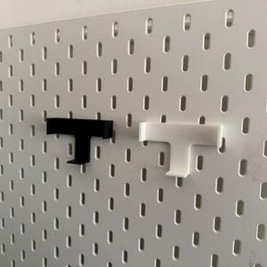 K&ouml;nnte beinhalten: Zwei T-f&ouml;rmige Lochwandhaken, einer schwarz und einer wei&szlig;, werden vor einer wei&szlig;en Lochwand gezeigt. Die Haken sind f&uuml;r die Verwendung an einer Lochwand konzipiert. Die Lochwand hat ein Gitter aus gleichm&auml;&szlig;ig verteilten L&ouml;chern.