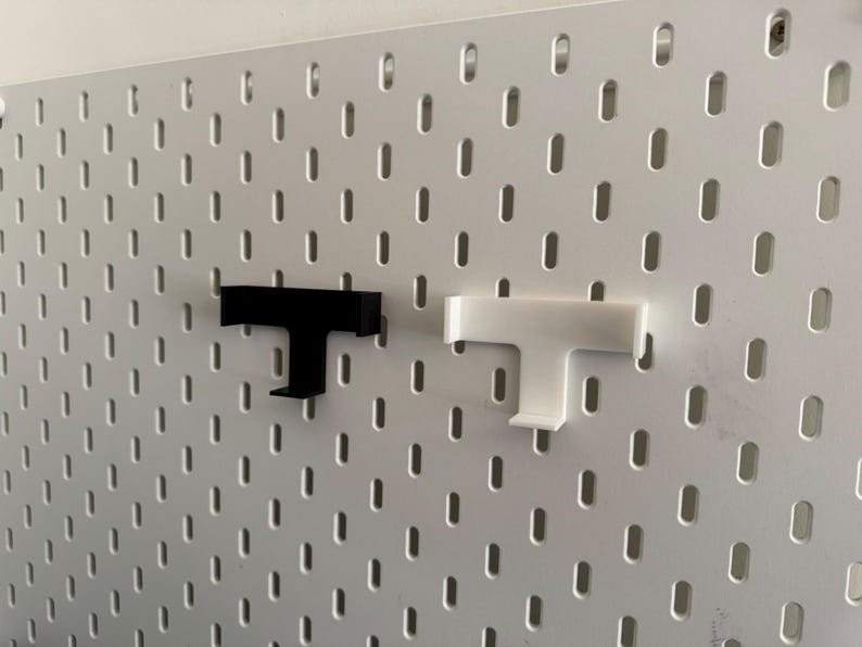 K&ouml;nnte beinhalten: Zwei T-f&ouml;rmige Lochwandhaken, einer schwarz und einer wei&szlig;, werden vor einer wei&szlig;en Lochwand gezeigt. Die Haken sind f&uuml;r die Verwendung an einer Lochwand konzipiert. Die Lochwand hat ein Gitter aus gleichm&auml;&szlig;ig verteilten L&ouml;chern.