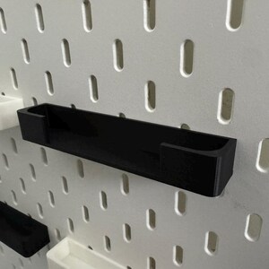 May include: Black rectangular pegboard shelf with a U-shaped design, mounted on a white pegboard. Additional white and black shelves are also visible. The shelves are designed for organising and storage.