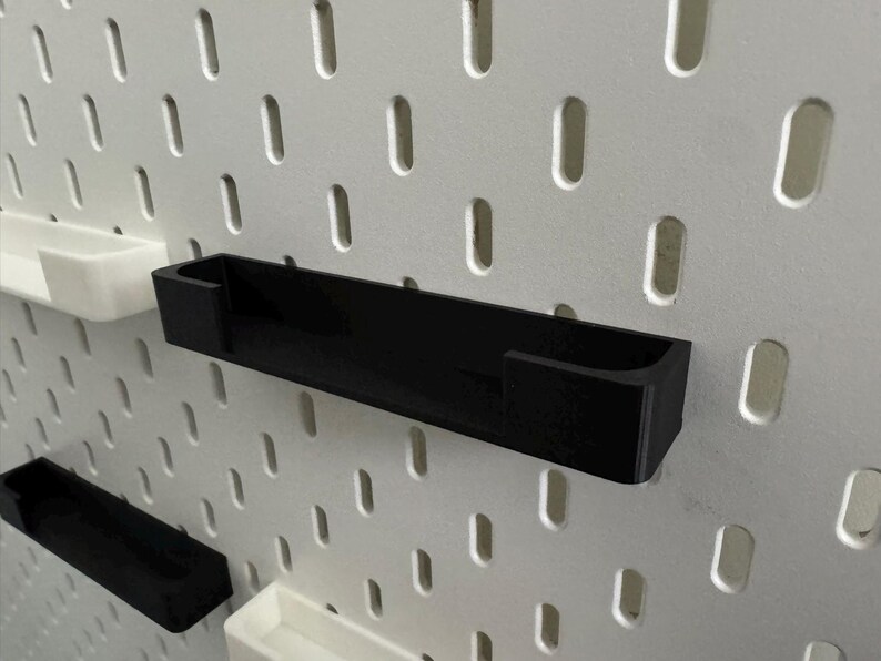 May include: Black rectangular pegboard shelf with a U-shaped design, mounted on a white pegboard. Additional white and black shelves are also visible. The shelves are designed for organising and storage.