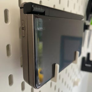 K&ouml;nnte beinhalten: Geschlossene, tragbare Dual-Screen-Spielkonsole in metallgrauer Ausf&uuml;hrung. Der obere Bildschirm ist schwarz, und die Konsole hat Lautst&auml;rkeregler an der Seite. Sie ist an einer wei&szlig;en Lochwand mit wei&szlig;en Haken befestigt.