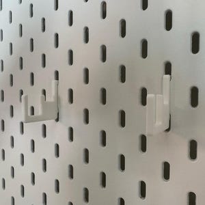 May include: White pegboard with evenly spaced oval holes. Two white plastic hooks are inserted into the pegboard. The hooks are designed to hold items. The pegboard is a versatile storage solution.