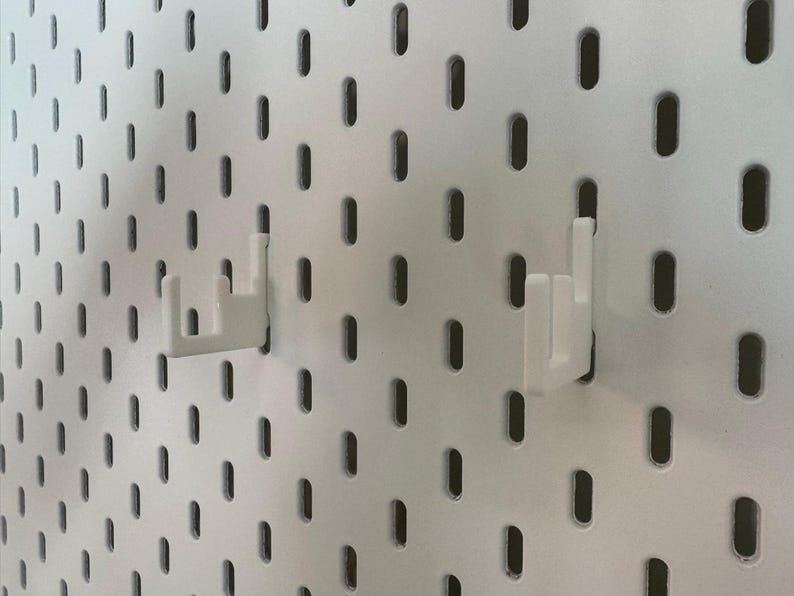 May include: White pegboard with evenly spaced oval holes. Two white plastic hooks are inserted into the pegboard. The hooks are designed to hold items. The pegboard is a versatile storage solution.