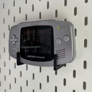 May include: A silver Game Boy Advance handheld console mounted on a black bracket against a white pegboard. The console has a screen, directional pad, and buttons. The text "GAME BOY ADVANCE" is visible.