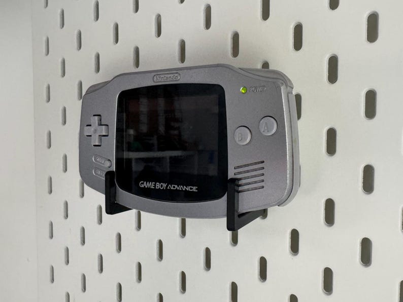 May include: A silver Game Boy Advance handheld console mounted on a black bracket against a white pegboard. The console has a screen, directional pad, and buttons. The text "GAME BOY ADVANCE" is visible.
