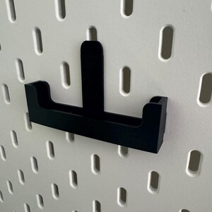 May include: Black plastic pegboard hook with a small shelf. The hook is designed to hold small items like tools or accessories.